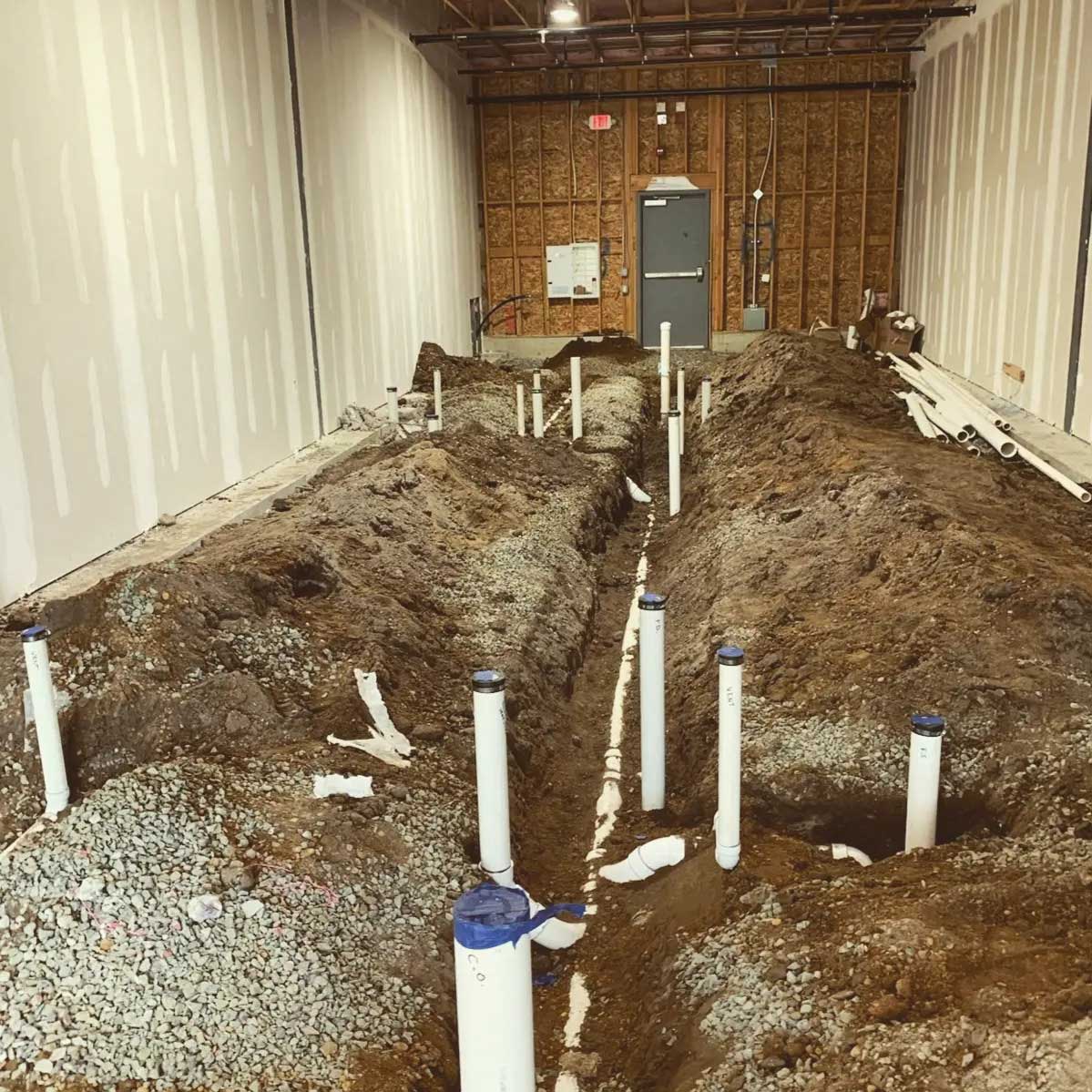 Indoor construction site with a large pile of dirt and gravel surrounded by white pipes sticking out of the ground, a wooden wall and a gray metal door in the background, and a concrete floor with scattered white pipes.
