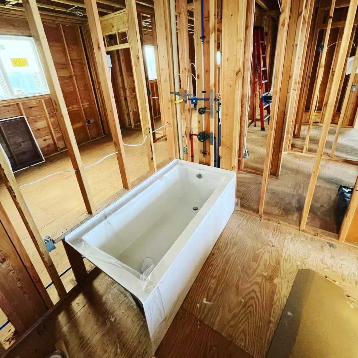 Bathroom under construction with wooden studs, plywood floor, white bathtub with blue cover and silver faucet, pipes and wires in walls and ceiling, window on left wall, and door on right wall.