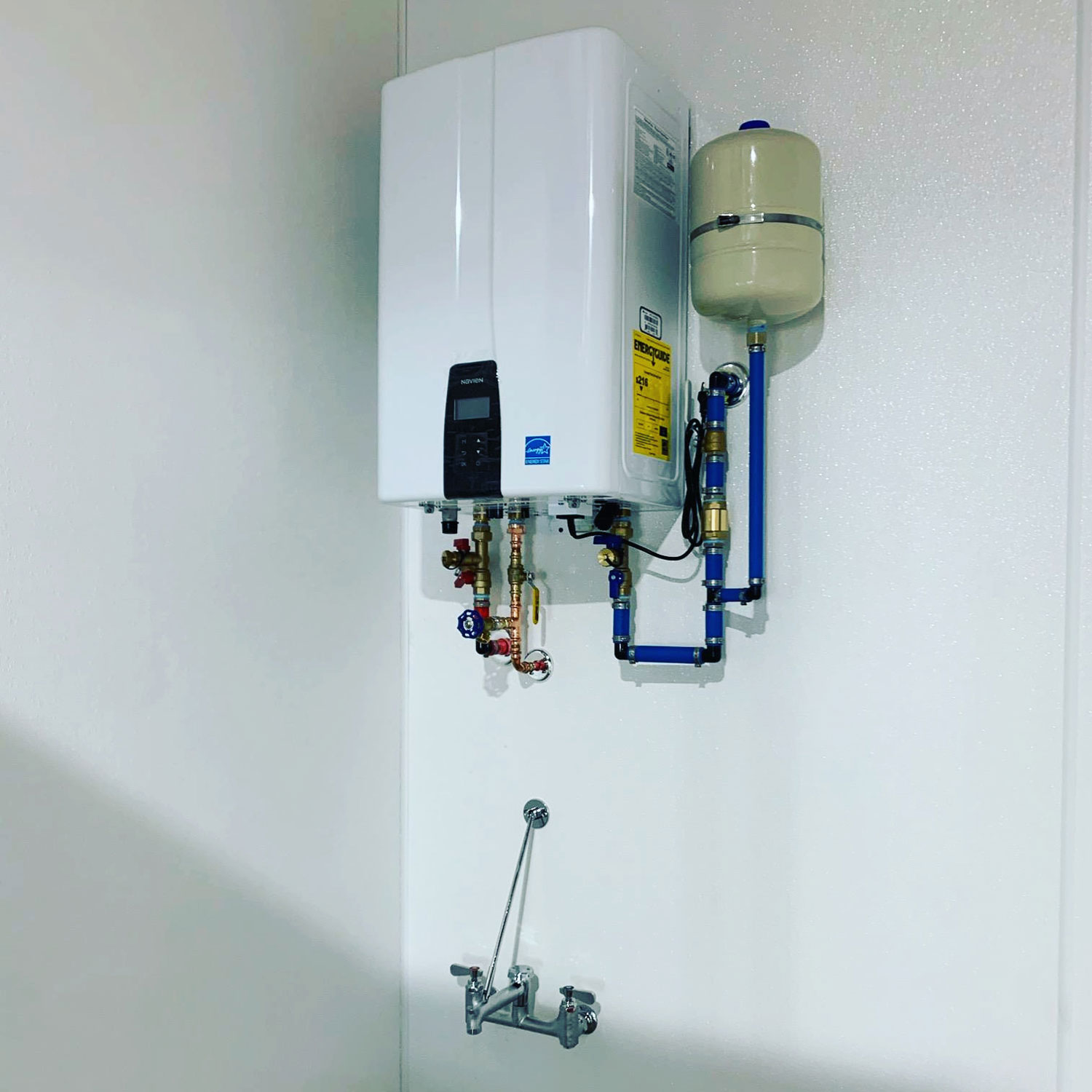 White tankless water heater with a digital display and a blue ‘Navien’ label, mounted on a white wall and connected to a blue and red piping system with multiple valves, and a silver faucet with a hose attached below.