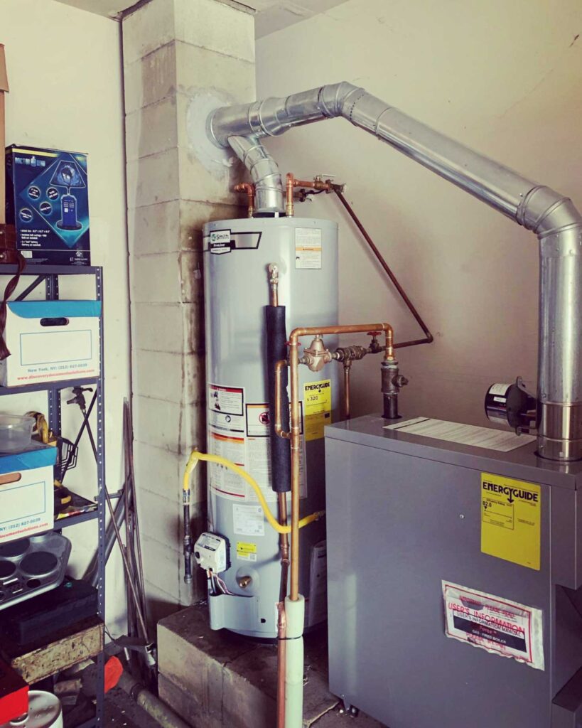 Utility room with a tall cylindrical water heater connected to a yellow gas line and a gray ‘energycube’ furnace, surrounded by pipes and ducts, against a concrete wall with a tool shelf.