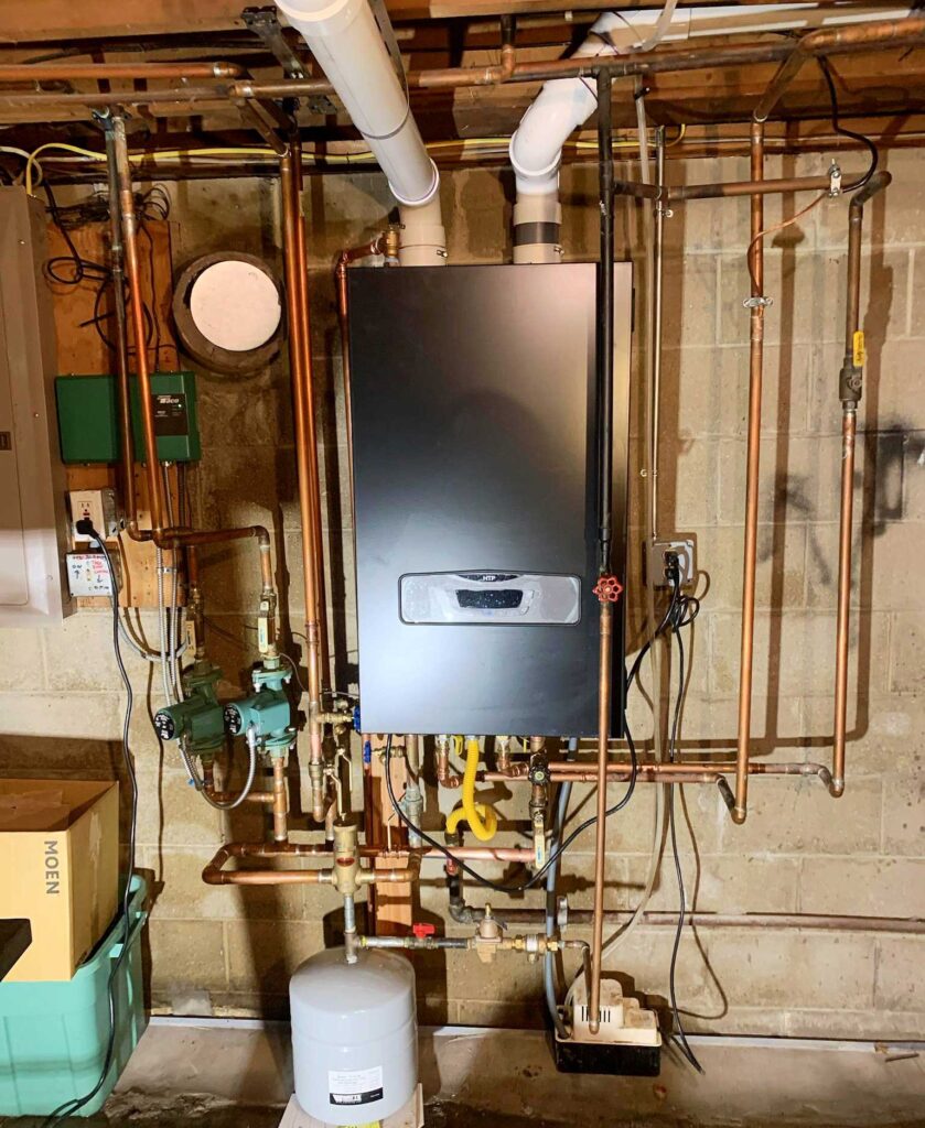 Large gray boiler in a boiler room, connected to copper and white pipes, with a green pump on the left and a white bucket on the right, against a concrete wall and wooden beam background.